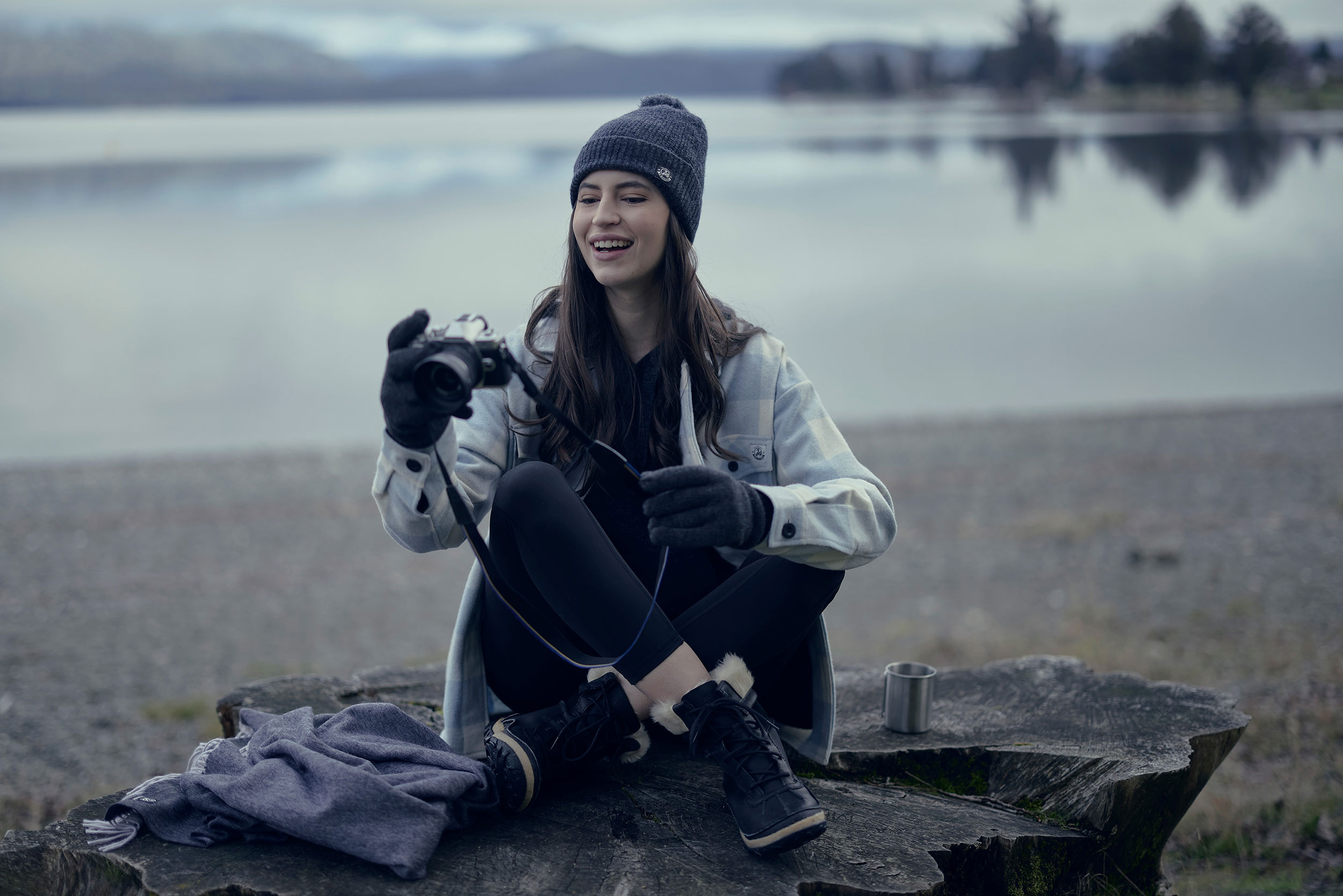 Smiling woman in winterwear with camera on picnic blanket by lakeside Winter